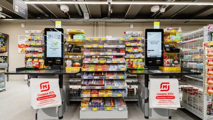 The image depicts a well-stocked convenience store with a modern design. The store features a large display of snacks and candies, neatly arranged on shelves and counters. Two self-service checkout stations are prominently displayed, each equipped with a touch screen and a cashier. The checkout counters are black and h