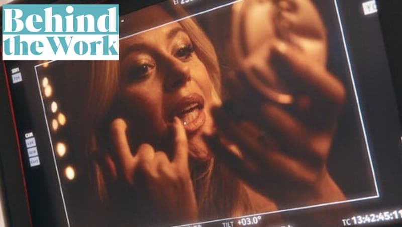 The image shows a woman with blonde hair, wearing a dark-colored top, holding a mirror and making a hand gesture. The scene appears to be set in a dark room, with a screen displaying a close-up of the woman's face. The screen is part of a video production setup, as indicated by the visible control panel on the left sid