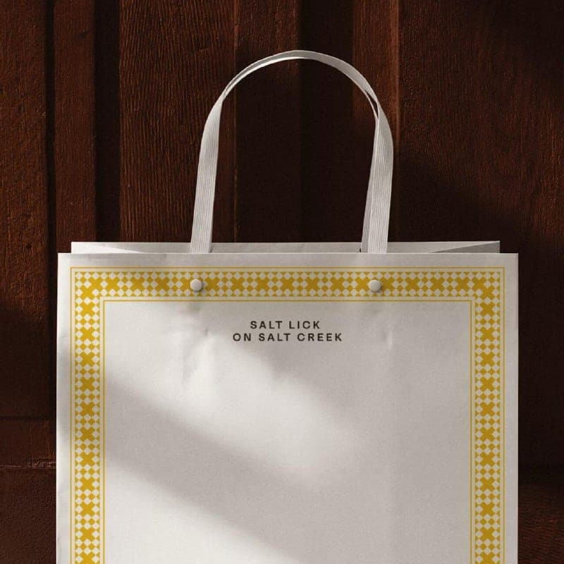 The image shows a white paper shopping bag with a vibrant yellow border featuring a repeating geometric pattern. The bag is positioned against a dark brown wooden wall, which provides a stark contrast to the bag's colors. The bag has a white handle and is slightly tilted to the right. The bag is empty, with no visible