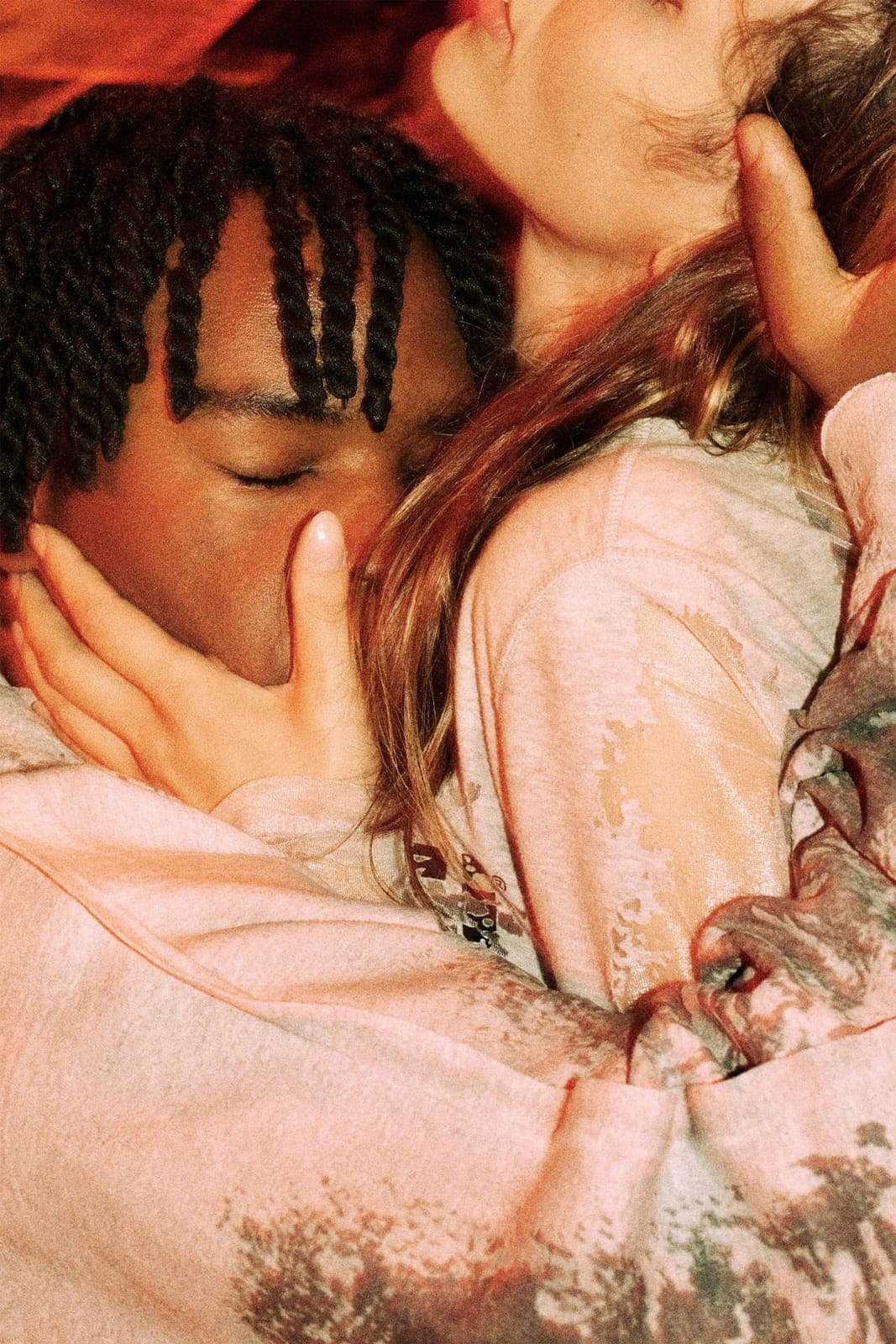 The image shows two individuals sharing a tender moment. The person on the left has dark skin and long, dark braids, and is wearing a light pink sweatshirt with a floral pattern. Their eyes are closed, and their head is resting on their hand. The person on the right has light skin and long, light brown hair. They are w