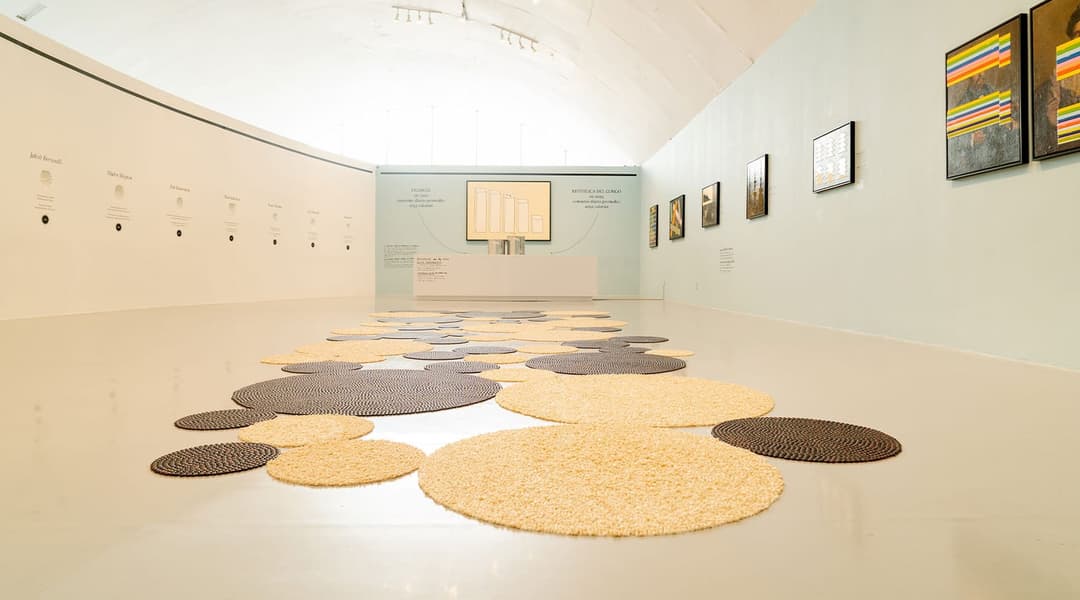 The image depicts an art gallery with a high ceiling and a white, curved ceiling. The gallery features a light beige or cream-colored floor and walls, with a light blue or teal accent wall. The walls are adorned with a series of framed artworks, including paintings and photographs, arranged in a linear fashion. A large