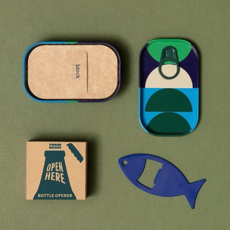 The image presents a collection of objects arranged on a green background. On the left, there is a brown cardboard box with a blue lid, featuring a design of a bottle opener and the text "Tinned Goods" and "Open Here". In the center, there is a blue tin can with a green lid, featuring a design of two overlapping circle