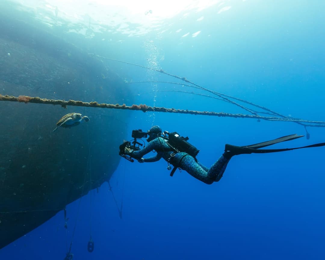 A scuba diver in a black wetsuit and black fins is captured in a moment of exploration in the deep blue ocean. The diver is holding a camera, poised to document the scene. A large, dark-colored object, possibly a ship or part of a ship, is visible in the background. A rope or cable is attached to the object, and a smal