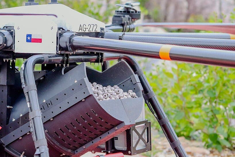 The image shows a drone equipped with a black basket attached to its side. The basket is filled with small, light-colored objects, possibly seeds or grains. The drone has a white body with black accents and a Texas flag on the side. The drone's body is primarily black with some red and orange accents. The drone is posi