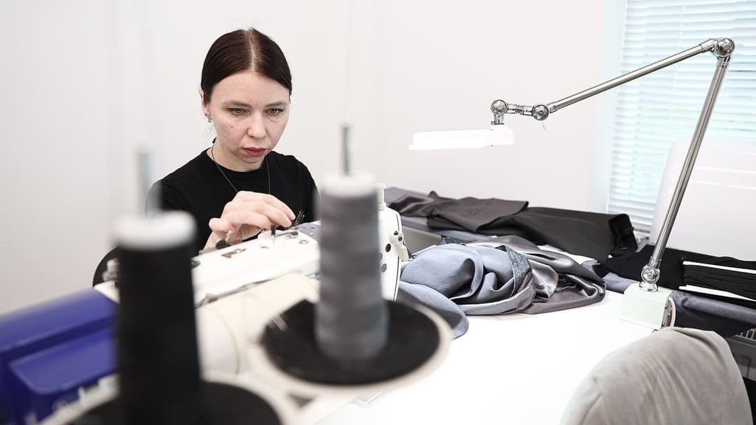 A woman with dark brown hair pulled back in a ponytail is seated at a white desk, engrossed in her work. She is wearing a black shirt and is using a sewing machine, which is positioned on the desk. The sewing machine is surrounded by spools of thread, and a gray fabric is visible on the desk. A metal lamp with a flexib