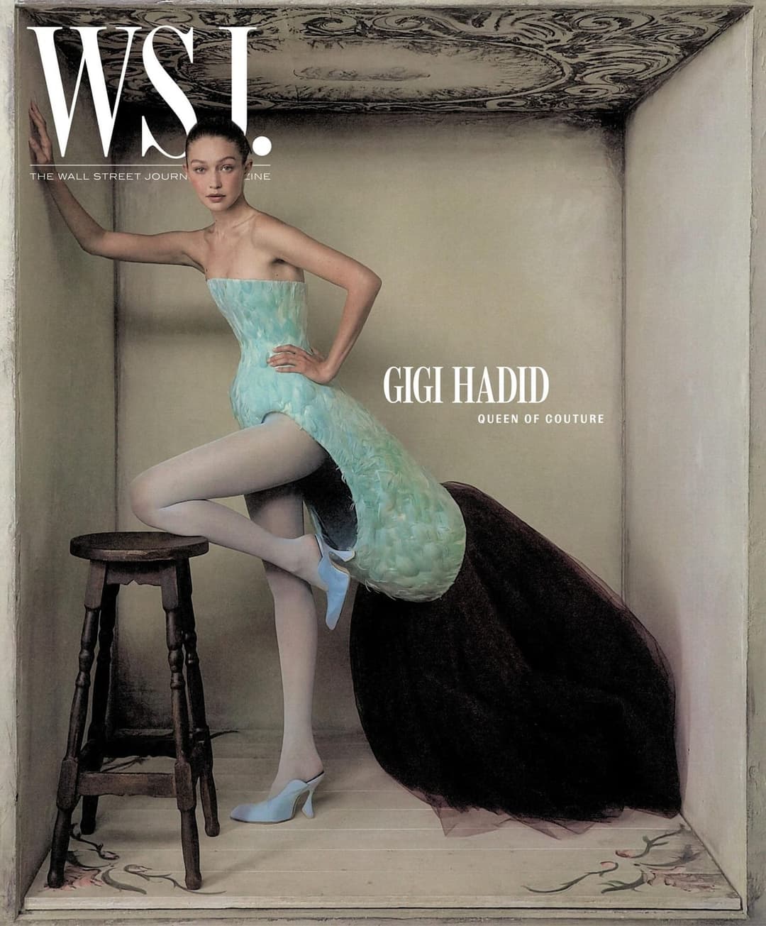 The image is a magazine cover for WSL, featuring Gigi Hadid. She is elegantly dressed in a light blue dress with a floral pattern and white tights, paired with light blue heels. She is standing in a small, white-walled room with a dark brown wooden stool in the foreground. The room has a decorative, possibly painted, c