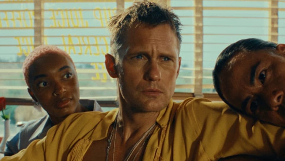 The image shows a man with short, light brown hair and a serious expression, wearing a vibrant yellow shirt. He is seated in front of a window with blinds, and his gaze is directed off to the side. Behind him, two other individuals are partially visible. One person has dark skin and short, light pink hair, and the othe