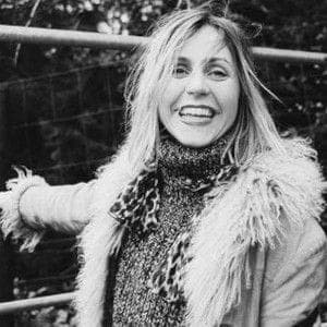 The image shows a young woman standing in front of a metal fence. She is wearing a light-colored jacket with a fur collar and a dark-colored, possibly knitted, sweater underneath. A scarf with a leopard print pattern is draped around her neck. Her blonde hair is slightly tousled, and she is smiling, looking directly at