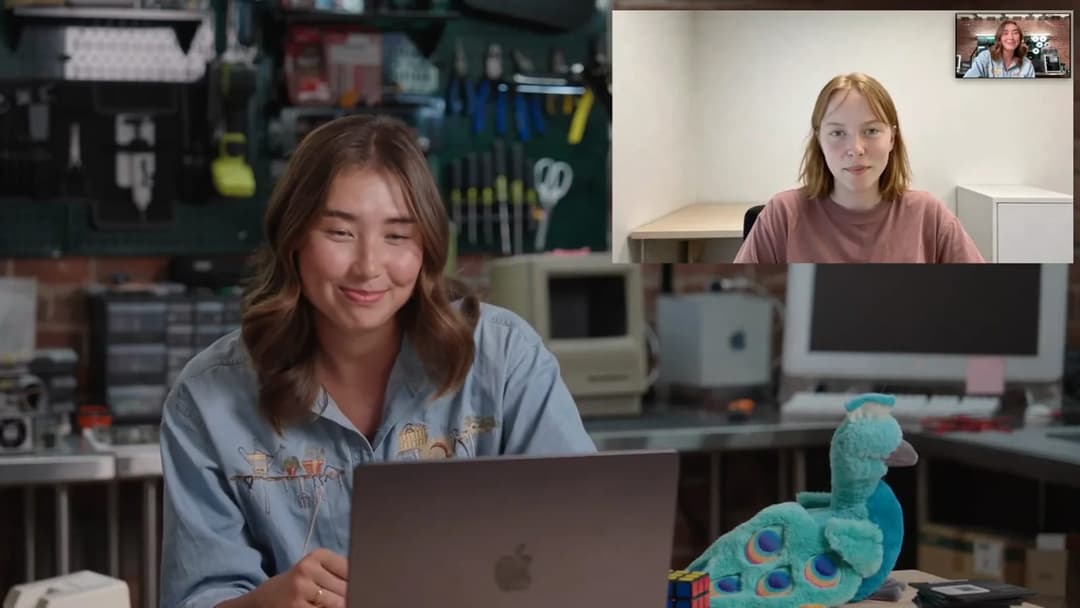 The image shows a young woman with long brown hair seated at a desk in a workshop or garage-like setting. She is wearing a light blue shirt with a design on the front and is smiling at the camera. A laptop is open on the desk in front of her. A peacock stuffed animal is also on the desk. The background features a clutt