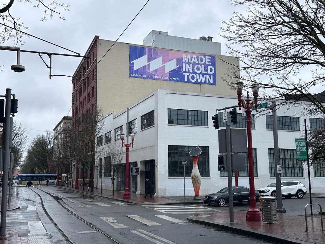 The image captures a city street scene on an overcast day. A large billboard on the side of a white building displays the text "MADE IN OLD TOWN" in bold, black letters. The building also has a sign that reads "INNOVATION IN FOOTWEAR AND APPAREL". The street is lined with trees, and a few cars are parked along the side