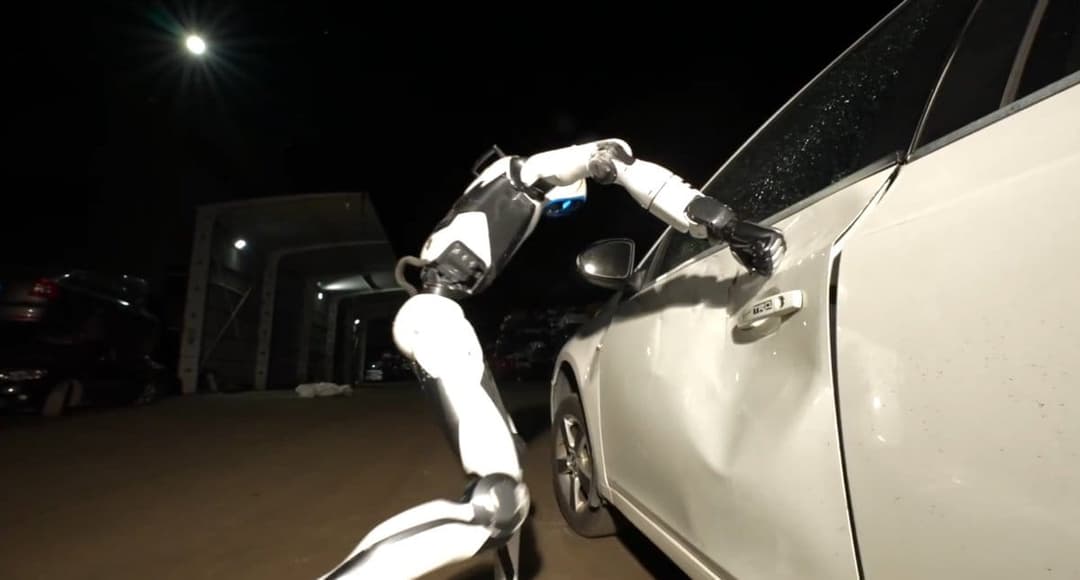 The image shows a robot with a white and black color scheme, featuring a white body and black arms and legs, interacting with a white car parked in a dimly lit parking lot. The robot's head is tilted towards the car's front door, and its hands are positioned on the door, possibly holding or adjusting something. The car