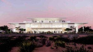 A rendering of a large white multi-tier building with a colonnaded front and glass facade under a pink dusk sky, with palm trees and desert-style landscaping in the foreground. No visible text.