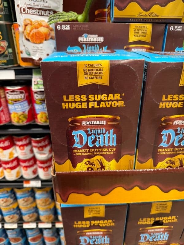 The image shows a display of Peanut Butter Cups from the brand "Feastables" stacked on a shelf in a grocery store. The cups are predominantly brown with a blue and gold design, and feature a chocolate drizzle pattern. The text on the cups reads "Less Sugar. Huge Flavor." and "Peanut Butter Cup - Soda-flavored Sparkling