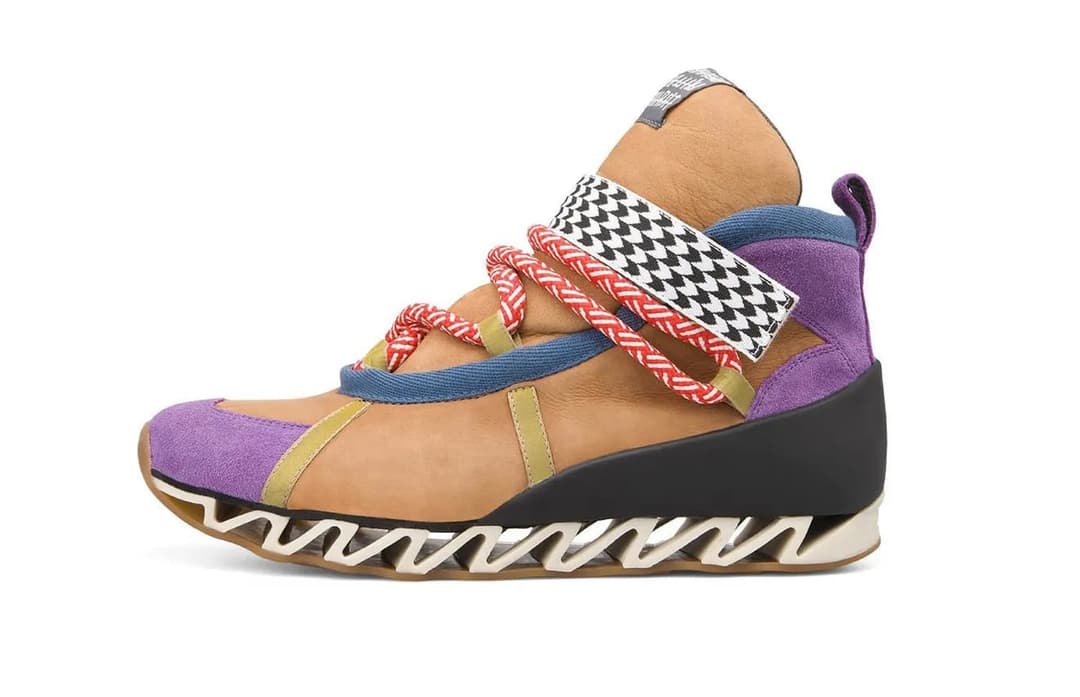 The image shows a single, high-top sneaker with a vibrant color scheme. The sneaker is primarily a tan or beige color, with a contrasting purple section on the side. The toe and heel areas are a dark blue or navy color. The midsole is a light beige or cream color with a unique, zigzag-like design. The laces are a mix o