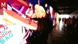 The image shows a person standing in a brightly lit hallway, holding a large, orange object that appears to be a giant ice cream cone. The hallway is illuminated by a series of colorful, illuminated screens, creating a vibrant atmosphere. The person is wearing a black jacket and has a large, blonde wig. The hallway is