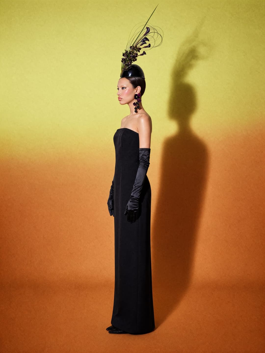 The image shows a woman standing against a vibrant yellow-to-orange gradient backdrop. She is elegantly dressed in a long black dress and black gloves, and her hair is styled in an updo with a black feather accessory. She is also wearing dark-colored earrings. The woman's shadow is cast on the backdrop, adding depth to