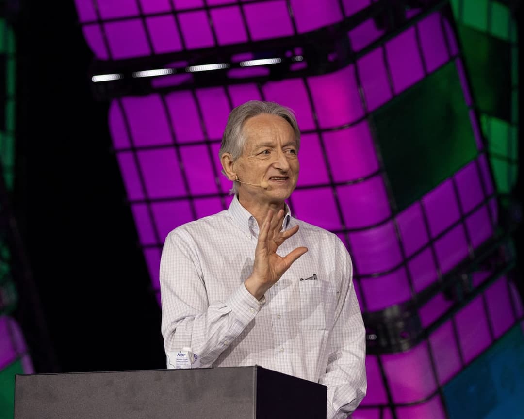The image shows a man standing at a podium, gesturing with his right hand. He is dressed in a white shirt with a subtle check pattern and has a microphone clipped to his shirt. The podium is positioned in front of a large screen displaying a vibrant purple and green geometric pattern. The man has gray hair and is weari