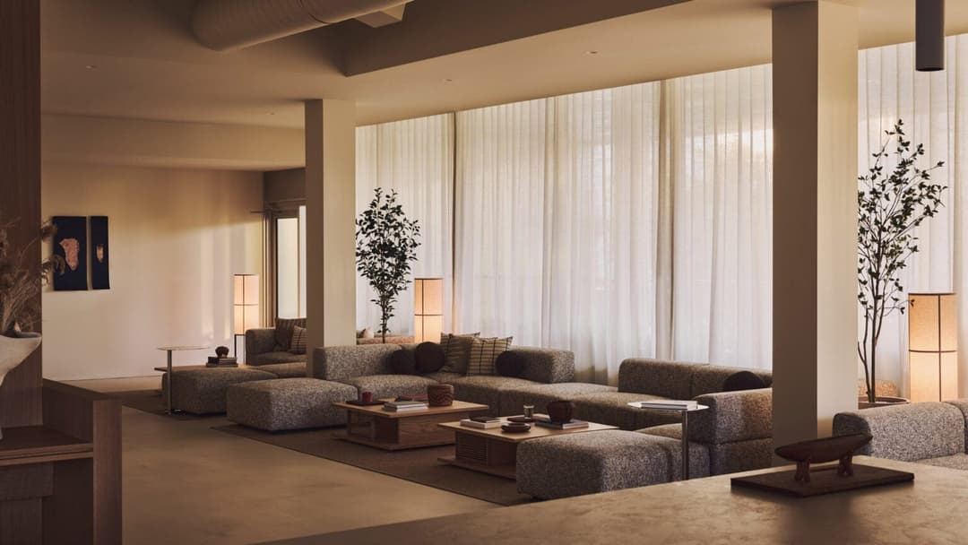 The image depicts a modern living room with a large window draped in white curtains, allowing natural light to fill the space. The room features a gray sofa and a matching gray armchair, arranged in a loose, inviting layout. A wooden coffee table sits in the center of the seating area, with a small side table beside it