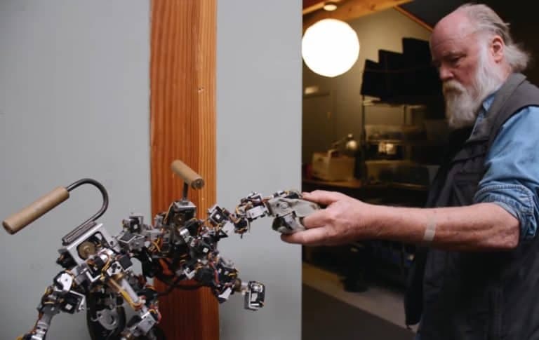 The image shows a man with a long white beard and gray hair, wearing a dark vest over a blue shirt, standing in a workshop. He is holding a small robotic arm, which appears to be made of metal and has a wooden handle. The man is looking at the robotic arm, possibly inspecting or adjusting it. The workshop has a light g