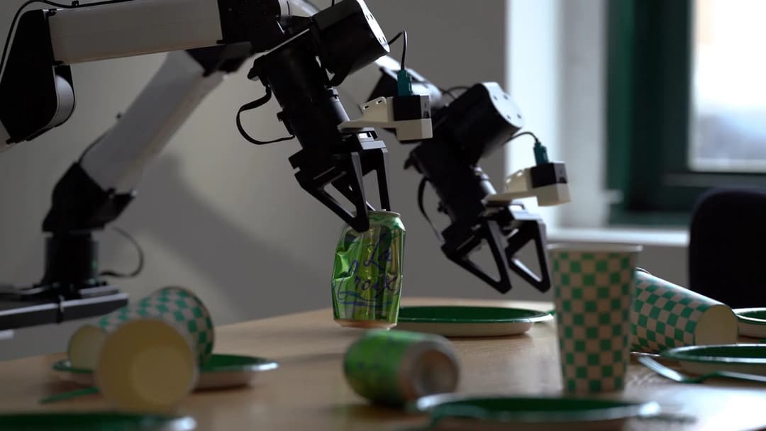 The image shows a black and white robotic arm reaching for a green soda can with a white label that reads "La Trox". The arm is positioned on a light brown wooden table, which also holds several green and white checkered paper cups and plates. The background features a window with a green frame, and a portion of a dark