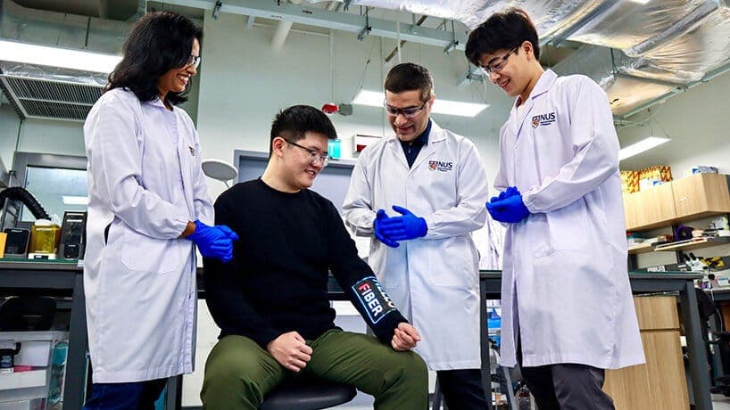 In a laboratory setting, four individuals are engaged in a scientific endeavor. The person in the center, wearing a black shirt and olive green pants, is seated and has a medical device attached to their forearm. The device is labeled "Fiber" and is connected to a blue wristband. Two individuals in white lab coats, one