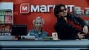 The image shows a man and a woman standing behind a checkout counter in a store. The man is wearing a black jacket and sunglasses, while the woman is wearing a gray shirt and has curly blonde hair. The store's red wall features the "MARKET" logo in white letters. The checkout counter is gray, and various items are visi