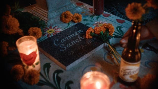 The image shows a table covered with a blue tablecloth featuring a floral pattern. On the table, there is a black stone plaque inscribed with "Carmen Sanchez" and "1899-2028". Two lit candles, one with a red rose design and the other unlit, are placed on the table. A small bottle of beer is also visible. Scattered arou