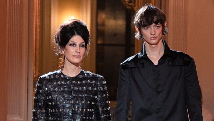 The image shows two individuals standing side by side in a room with ornate architectural details. The woman on the left is wearing a black jacket with a textured pattern, and her hair is styled in a classic updo. The man on the right is wearing a black shirt and has a casual, relaxed posture. The room features a large
