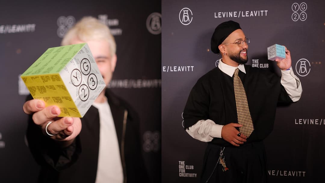 The image shows two individuals standing against a black backdrop adorned with white text and logos. The person on the left is holding a cube-shaped object with a green and yellow design, featuring the numbers "2", "3", and "Y" in a pattern. The person on the right is wearing a black suit, a white shirt, and a black be