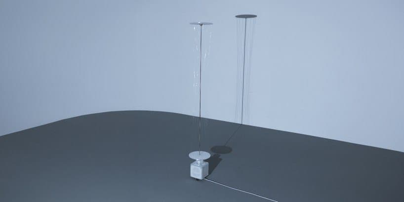 The image shows a minimalist scene with two tall, thin, silver-colored lamps standing on a dark gray table. The lamps have a circular base and a thin, vertical stem extending upwards. The lamps are positioned in the center of the image, casting shadows on the table. The background is a light blue color, providing a con