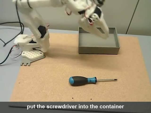 The image shows a white robotic arm with black and gray details, positioned on a light gray or beige surface. The arm is holding a gray box in its right hand and appears to be in the process of placing it into a gray container. A blue screwdriver with a black handle lies on the surface near the arm. The text "put the s