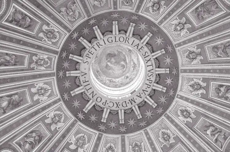 The image depicts the interior of a dome, possibly the St. Peter's Basilica in Vatican City, viewed from a low angle. The dome is adorned with intricate frescoes and paintings, including depictions of angels and celestial bodies, creating a sense of awe and grandeur. The frescoes are painted in shades of gray and white