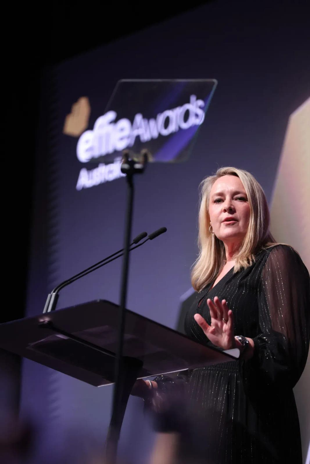 The image shows a woman standing at a podium, speaking into a microphone. She is dressed in a black dress with a subtle shimmer or sparkle pattern. The podium is positioned in front of a large screen displaying the words "Elite Awards Australia". The woman has blonde hair and is wearing a watch on her left wrist. The b