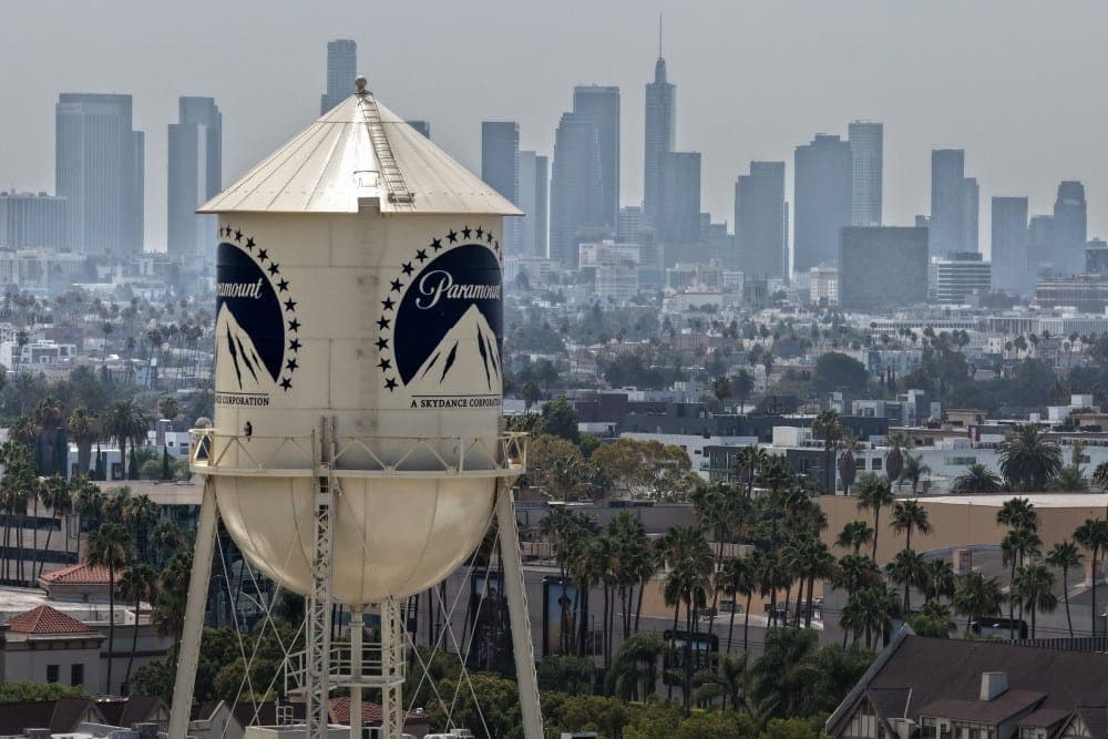 The image shows a large, white water tower with a curved roof and a ladder on top, standing prominently in the foreground. The water tower is adorned with the Paramount logo, a blue circle with a white mountain and the word "Paramount" written in white. The tower is surrounded by a lush expanse of palm trees, and the c