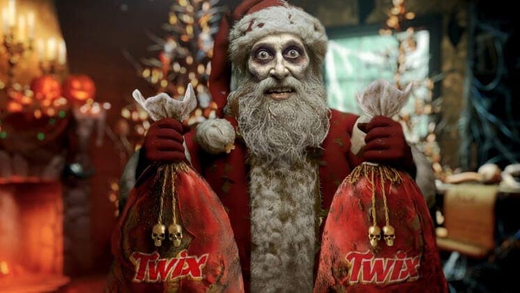 The image shows a figure resembling Santa Claus, dressed in a traditional red suit with a white beard and hat, holding two red Twix bags with gold handles. The figure's face is painted white with a creepy expression, and the bags are labeled with the word "Twix" in bold red letters. The background is out of focus, but