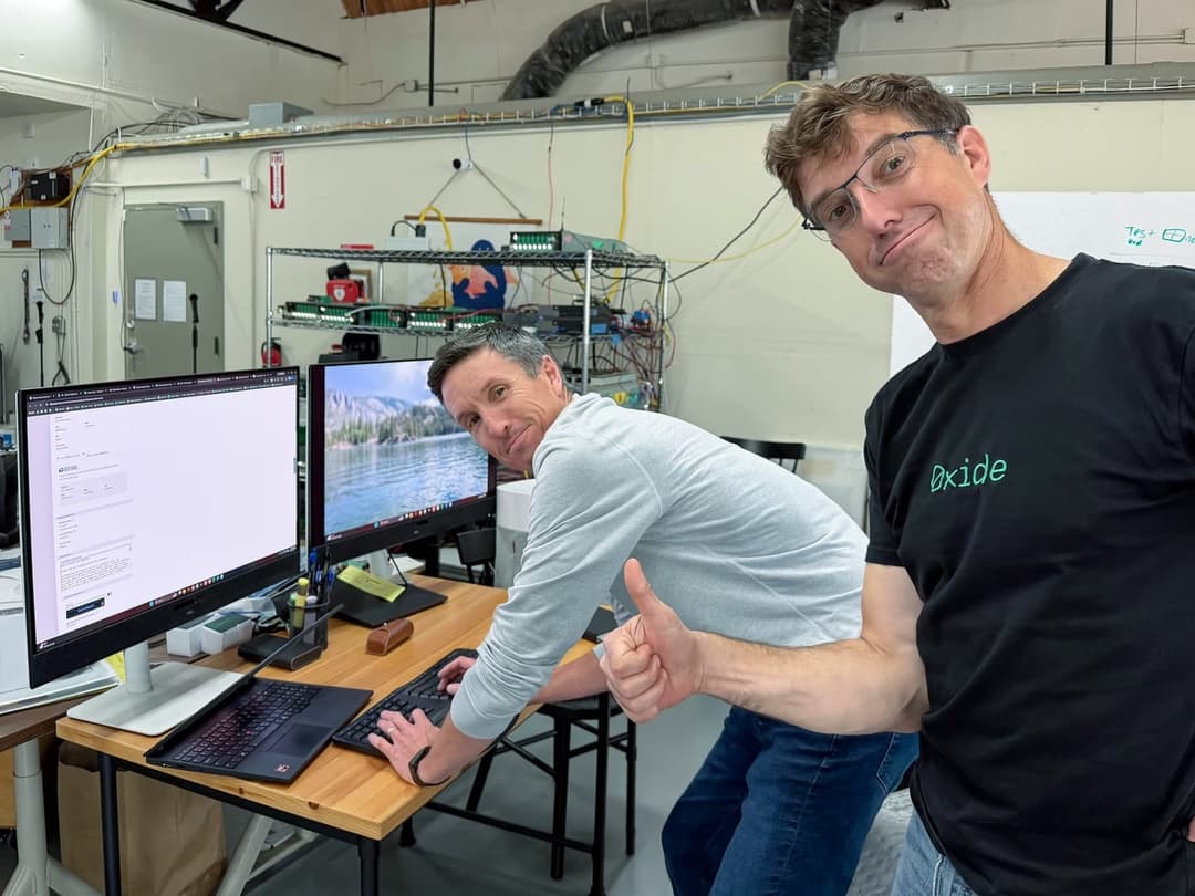 The image shows two men in a workshop setting. One man is wearing a black t-shirt with the word "Oxide" printed on it, and the other is wearing a light gray t-shirt. The man in the black t-shirt is giving a thumbs-up, while the man in the light gray t-shirt is leaning over a wooden desk, looking at a computer screen. T