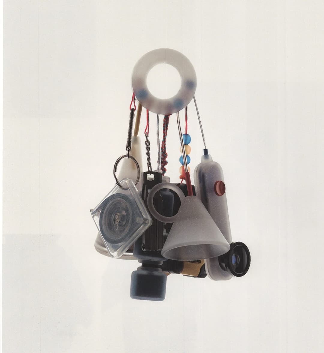 The image shows a collection of objects suspended from a white string against a white background. The objects include a round, translucent, light-colored ring, a small, dark-colored, possibly black, cylindrical object, a small, dark-colored, possibly black, cylindrical object, a small, dark-colored, possibly black, cyl