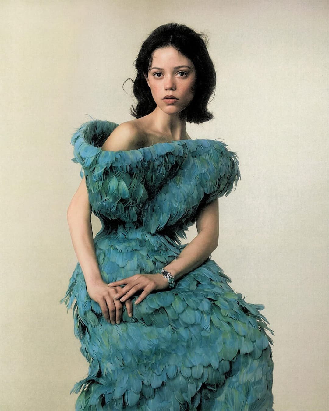 The image shows a young woman with dark hair and a serious expression, standing against a beige background. She is wearing a striking blue dress made of feathers, which drapes over her shoulders and falls to her waist. The dress has a loose, flowing fit and is adorned with a large, fluffy collar. She is also wearing a
