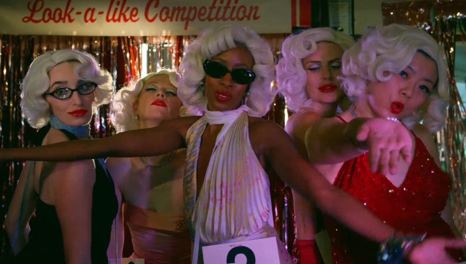 The image shows five women dressed in glamorous outfits, possibly from a film or television show, posing in front of a shimmering gold and red curtain. The women are wearing white wigs and sunglasses, and their outfits include a variety of colors and styles. One woman in the center is wearing a white dress with a numbe