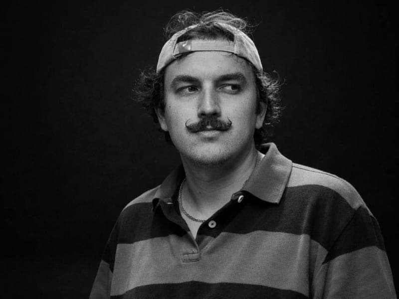 This black and white photograph captures a man with a mustache and a cap on his head. He is wearing a striped polo shirt and a necklace. The man's gaze is directed off to the side, and his expression is neutral. The background is a solid black, providing a stark contrast to the man's features and clothing.