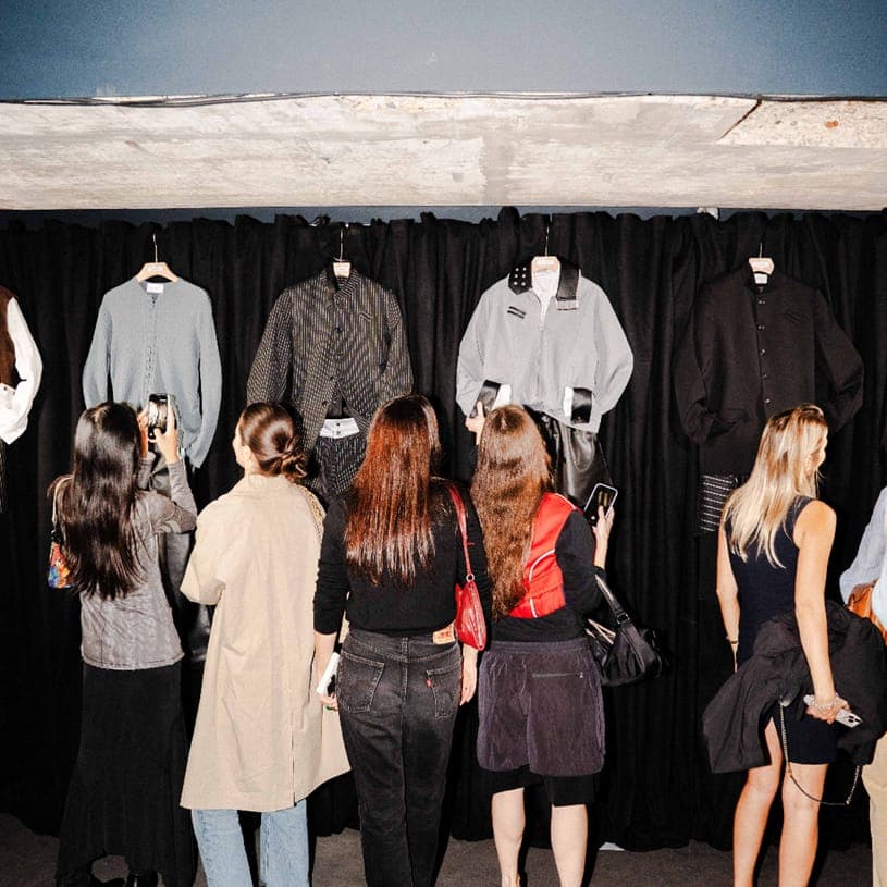 The image shows a group of people gathered in front of a black curtain, which serves as a backdrop for a clothing display. The display features a variety of clothing items, including a gray sweater, a black jacket, a gray shirt, a black jacket, a black shirt, a gray shirt, a black skirt, and a black jacket. The clothin