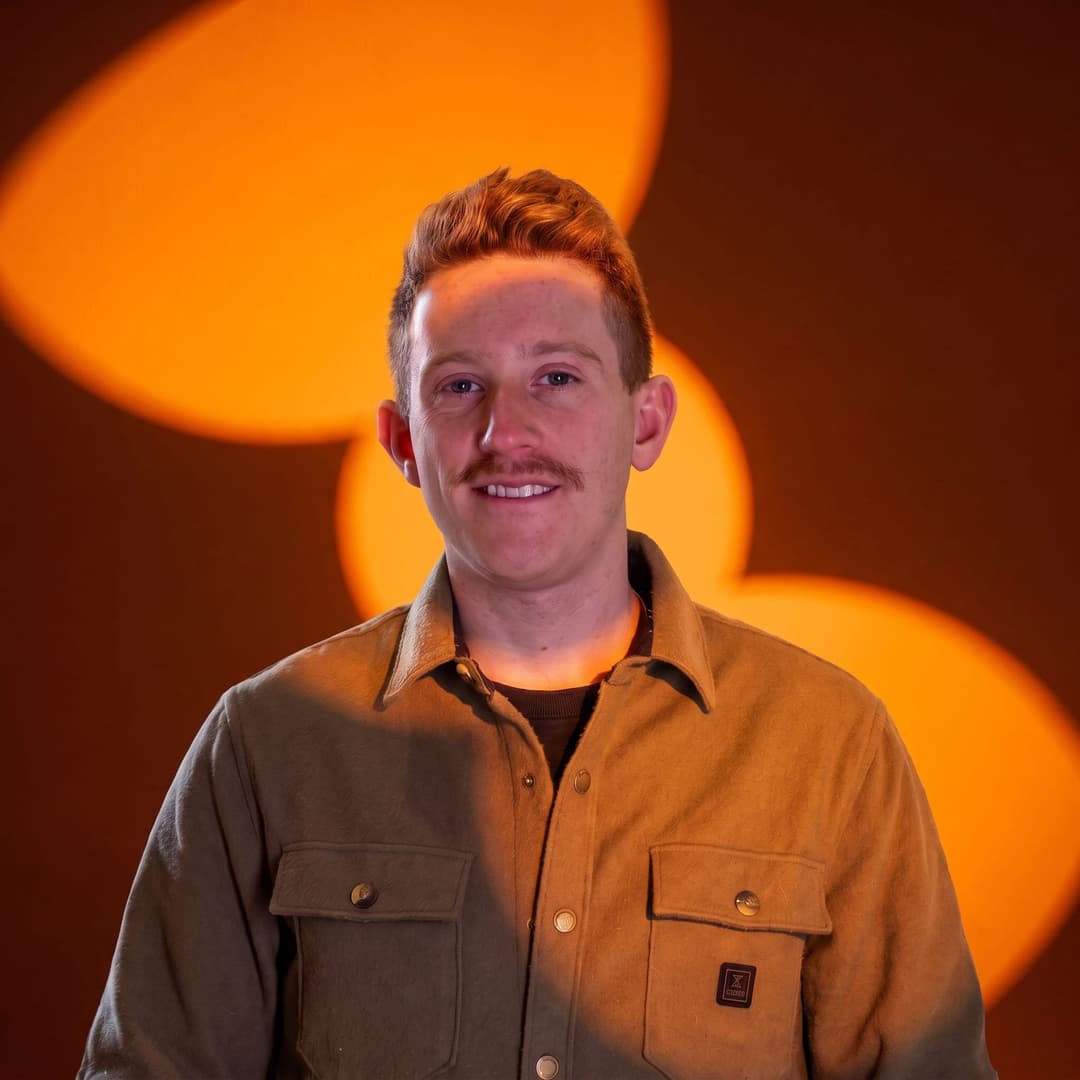 The image shows a young man with red hair and a mustache, wearing a brown button-down shirt with a small logo on the left chest pocket. He is standing against a backdrop of three large, orange-toned circles that cast a warm glow on his face. The man is looking directly at the camera with a slight smile.