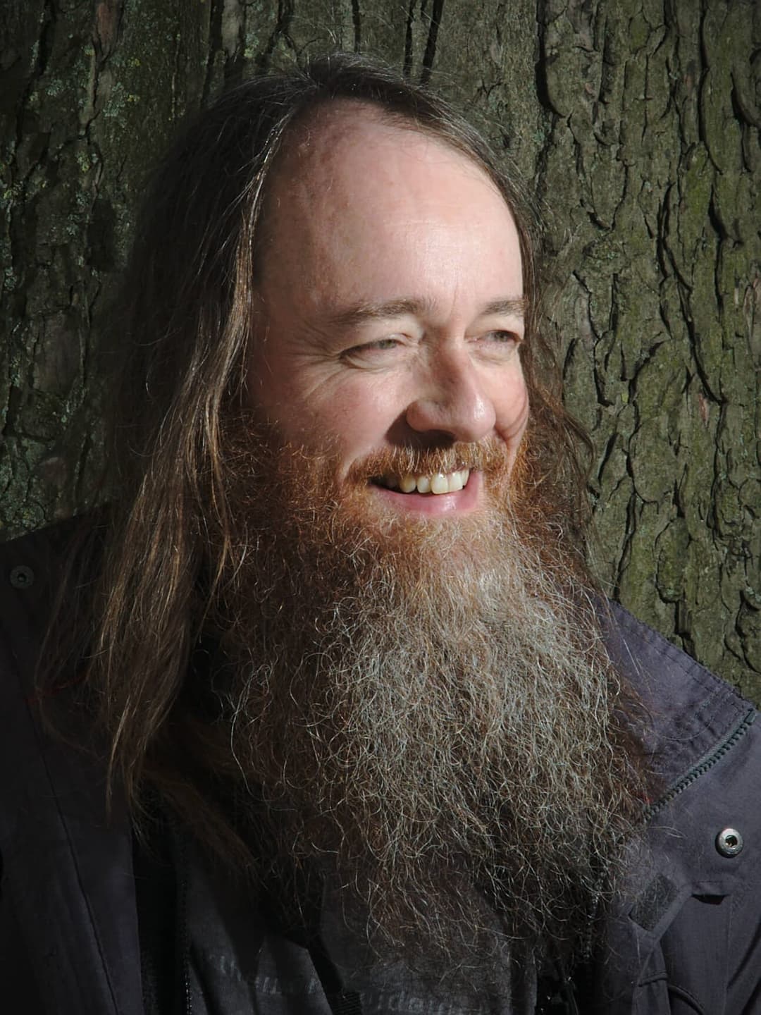 The image shows a man with long, dark brown hair and a full, gray beard. He is wearing a dark gray or black jacket and smiling warmly. The man is positioned in front of a tree trunk, which serves as a backdrop for the image. The tree trunk is covered in moss, adding a natural element to the scene. The man's gaze is dir