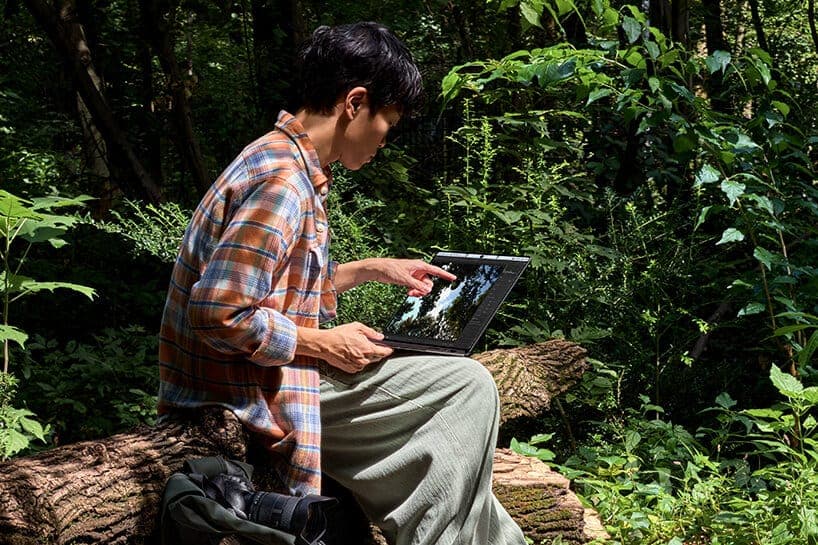 A person in a plaid shirt sits on a mossy log in a green forest and touches a laptop screen showing a photo of trees and sky. The scene is a lifestyle product shot.