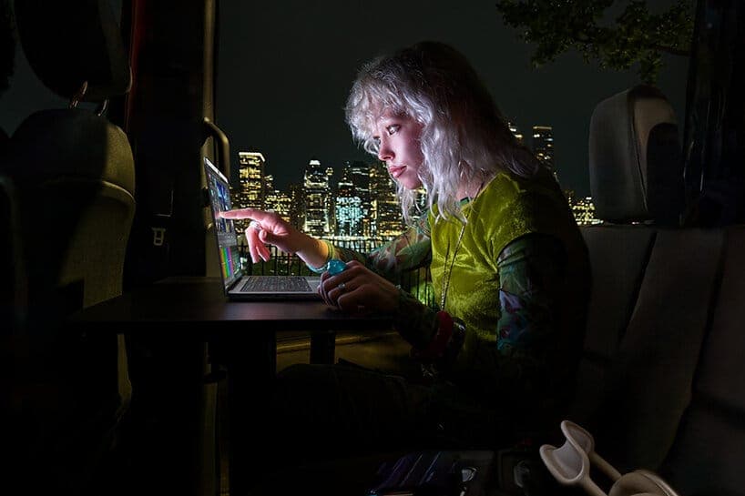 A woman with silver-white curly hair in a lime-green top points at a laptop screen in a dark vehicle at night, with an illuminated city skyline visible through the window. No legible brand text is visible.