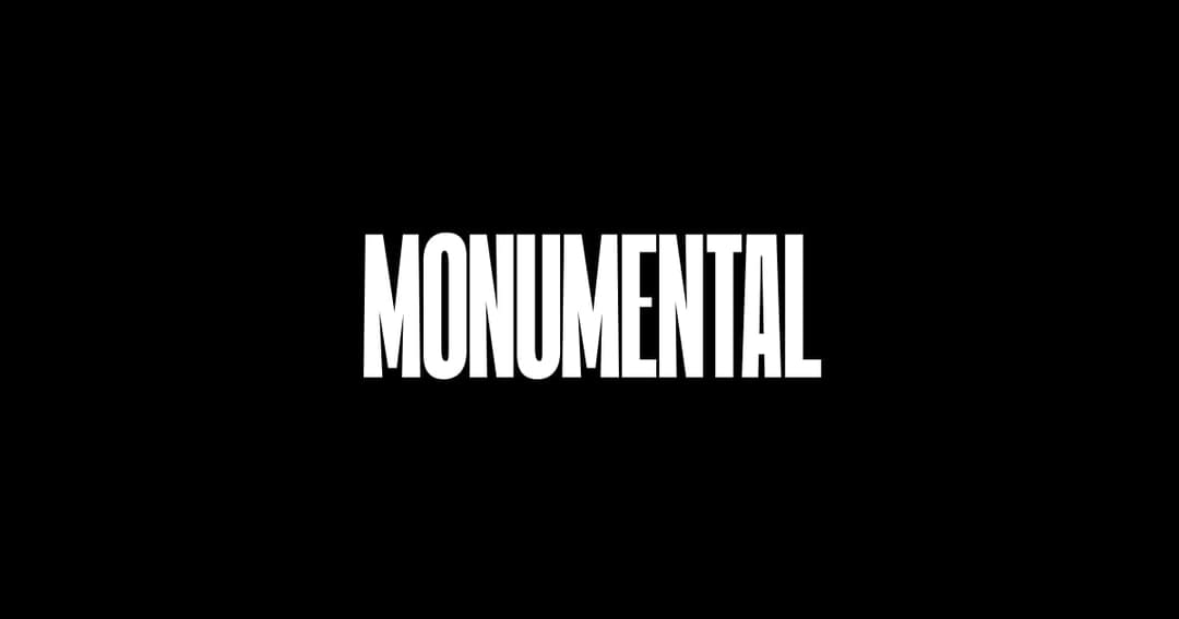 The image presents a stark contrast of black and white. Dominating the center of the image is the word "MONUMENTAL" written in bold, white, sans-serif capital letters. The text is centered and occupies the entire frame, creating a sense of balance and symmetry. The black background serves as a strong backdrop, making t