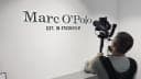 The image shows a man standing in front of a white wall, holding a black camera mounted on a tripod. The wall features a black and white logo that reads "Marc O'Polo" and "Est. in St. Louis". The man is wearing a gray t-shirt and a black camera strap. The setting appears to be a room with a light gray or white countert