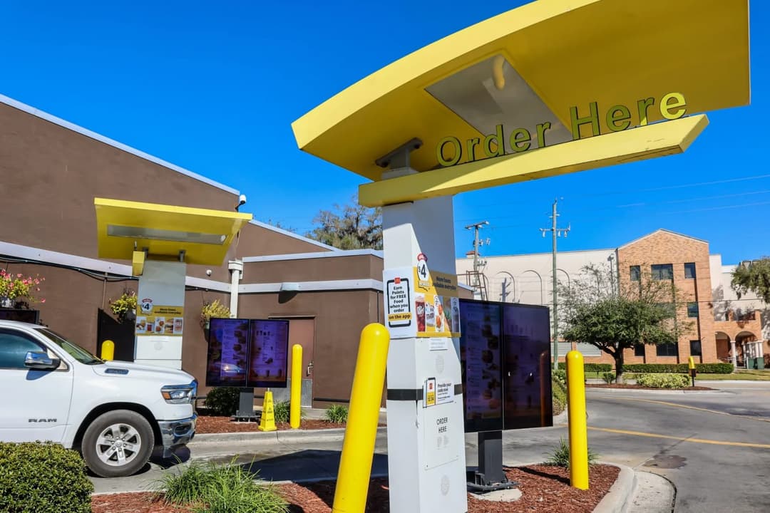 The image shows a McDonald's restaurant with a large yellow "Order Here" sign prominently displayed. The restaurant is situated in a parking lot, with a white pickup truck parked nearby. The parking lot is bordered by a curb and features yellow poles. The restaurant's exterior is primarily brown, with a section of a br