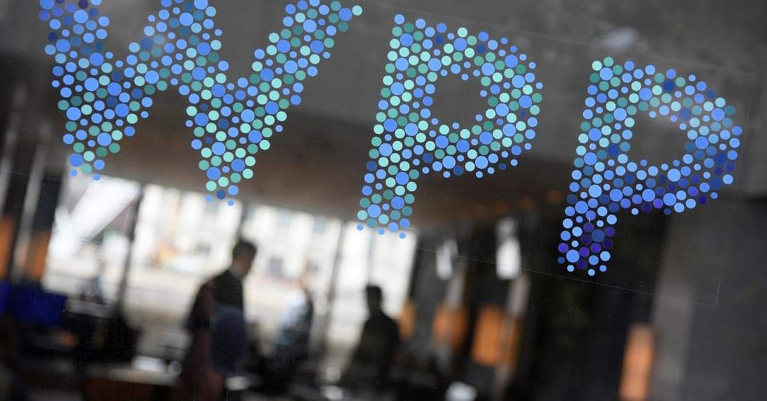 The image shows a glass window with a blue and teal dot pattern spelling out the letters "WPP". The dots are small and round, creating a textured appearance. The window reflects the blurred interior of a building, which appears to be an office or commercial space. The reflection also shows a few people walking in the b