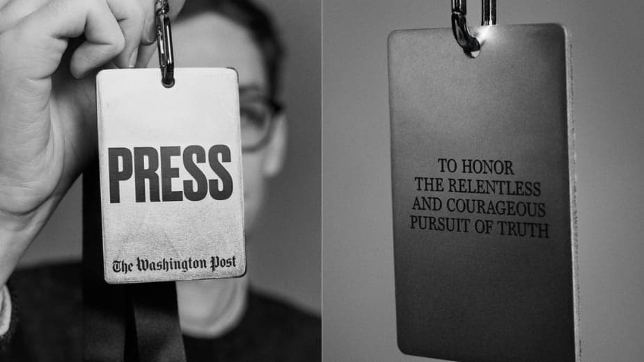 The image is a black and white photograph featuring a person holding a rectangular sign with a silver/grey background. The sign displays the word "PRESS" in bold, black letters, and the text "The Washington Post" is printed in smaller font below it. The person is wearing glasses and a dark-colored shirt. The sign is at